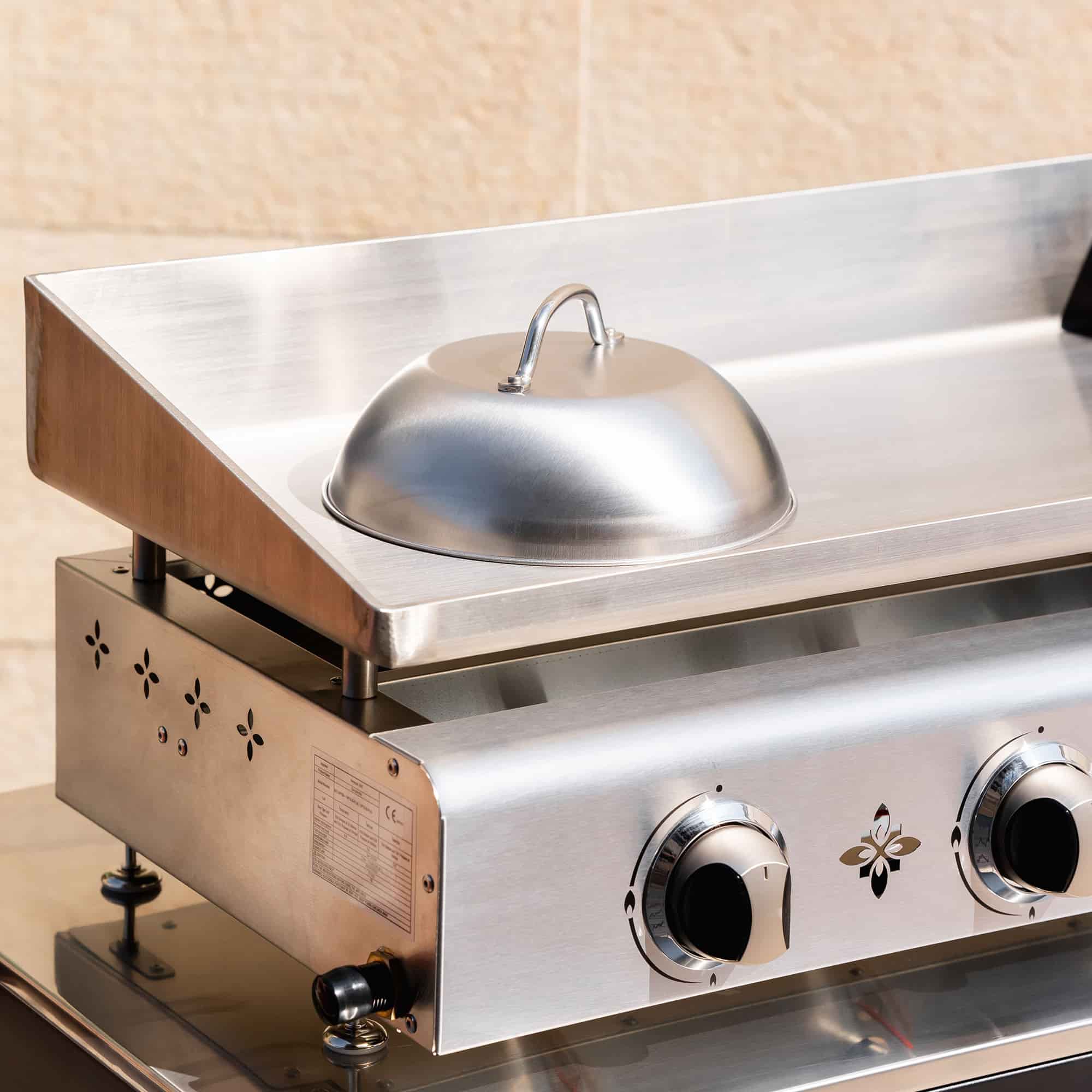 Cúpula de cozedura em inox pousada sobre a placa de uma plancha em inox de três queimadores, apresentada no exterior em grande plano.