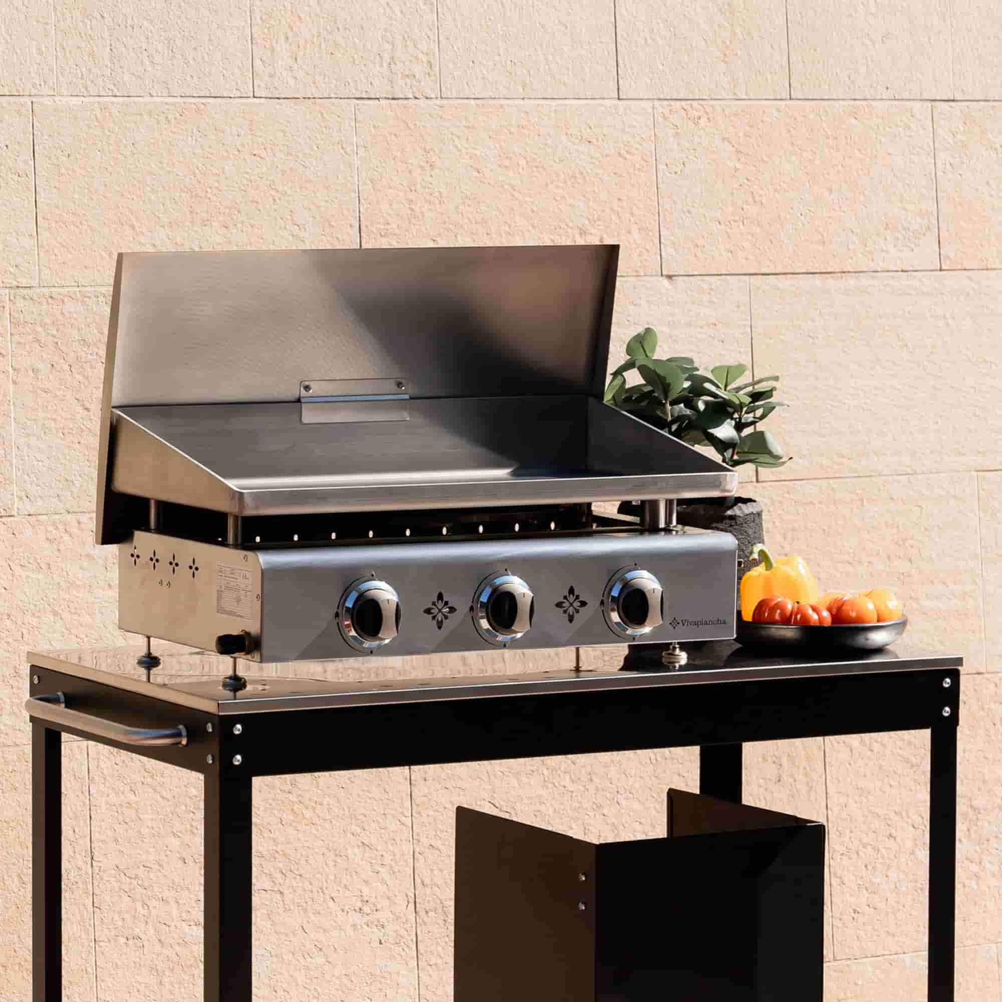 Plancha em inox com três queimadores e tampa levantada, colocada num carrinho preto em frente a uma parede clara, acompanhada de legumes e de uma planta decorativa.