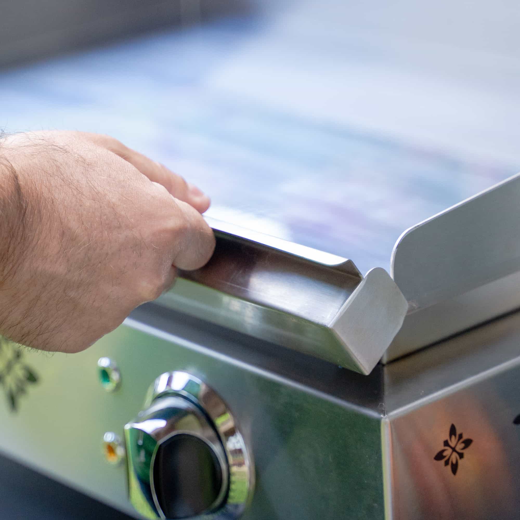 Grande plano da mão de uma pessoa a manusear o rebordo da placa em inox de uma plancha elétrica Vivaplancha, com o botão de regulação visível em primeiro plano.