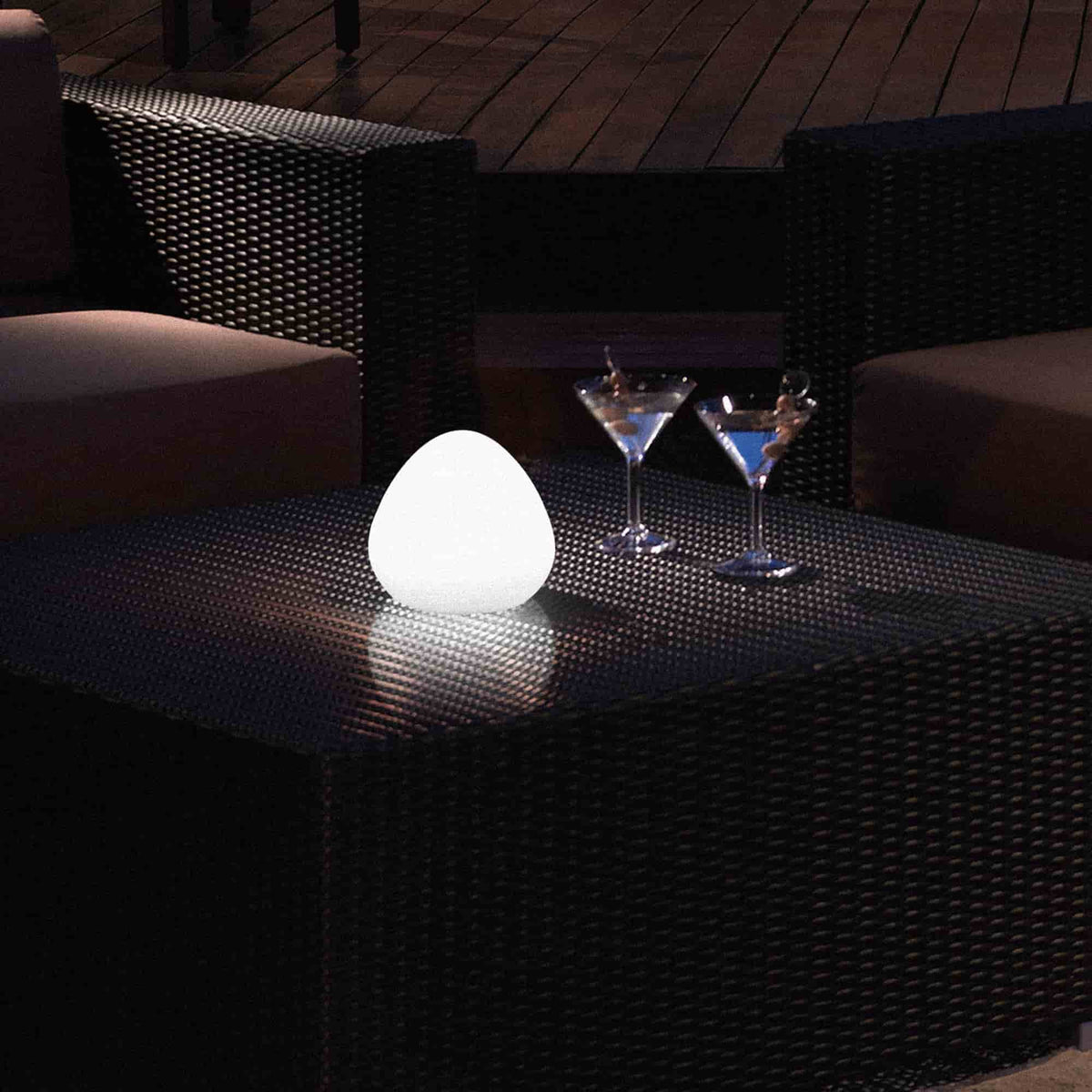 Lampe solaire d’extérieur en forme de noisette, posée sur une table en résine tressée, diffusant une lumière blanche douce dans une ambiance de terrasse nocturne.