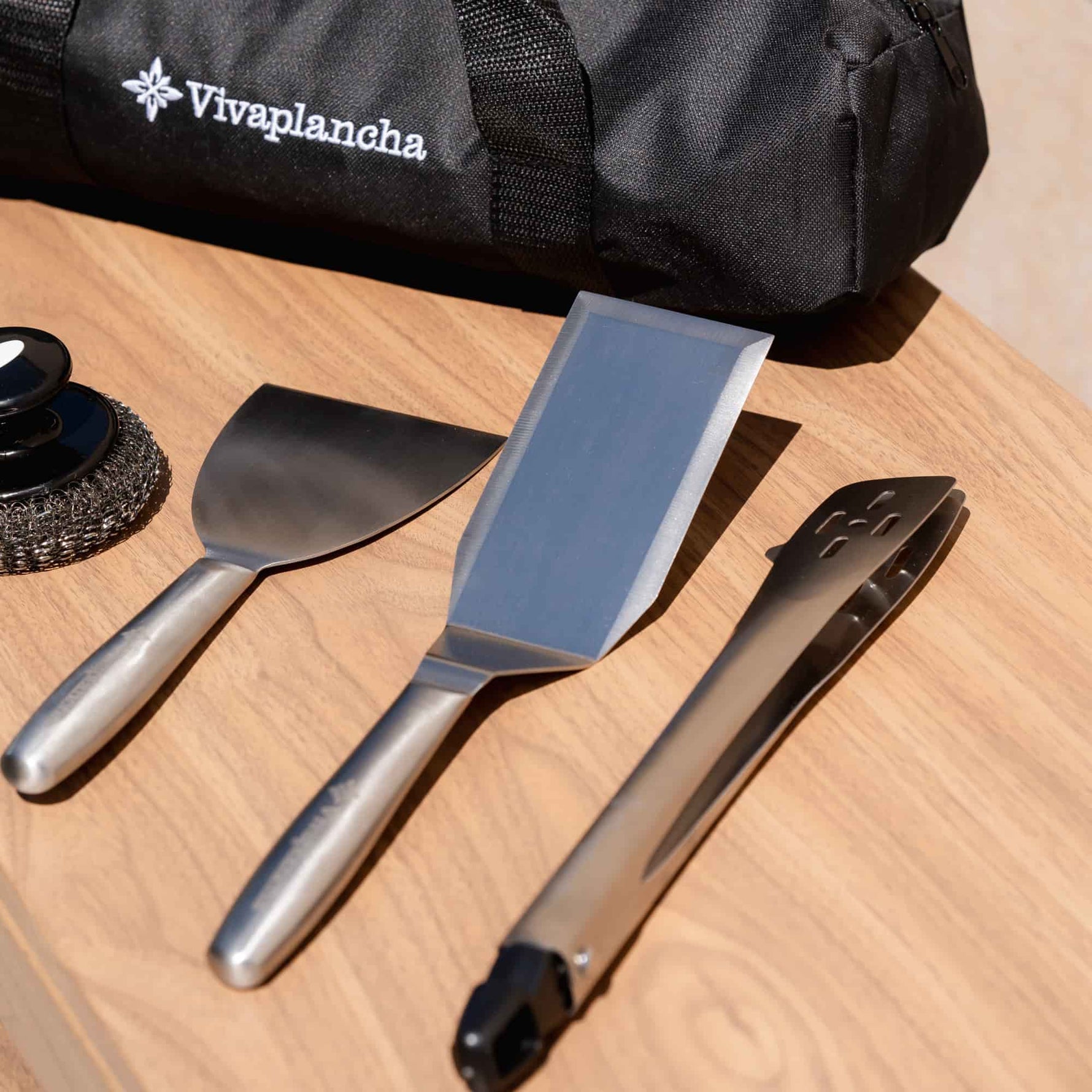 Ustensiles Vivaplancha posés sur un plan de travail en bois : spatule large, spatule longue, pince en inox et boule de nettoyage, avec le sac de transport visible en arrière-plan.