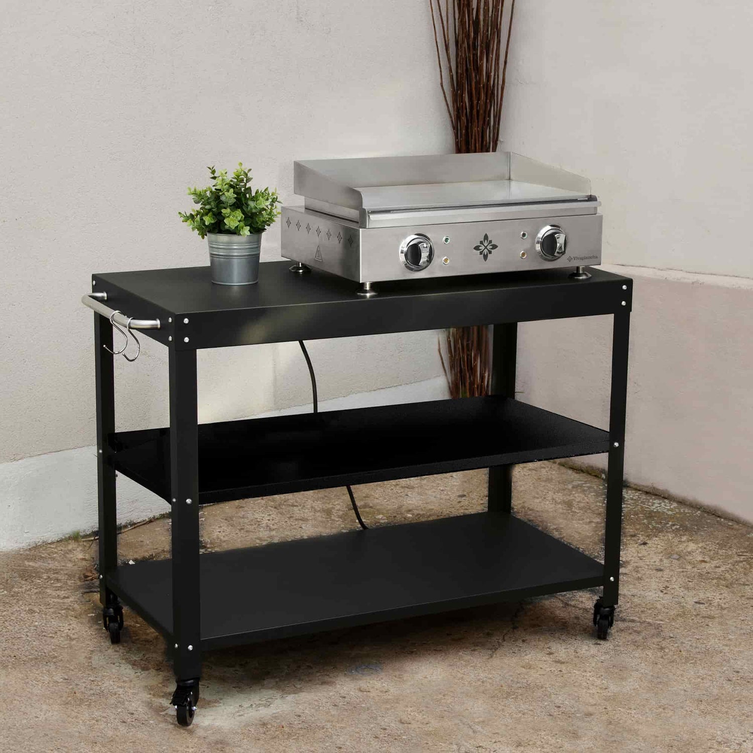 Chariot noir pour plancha avec trois niveaux de rangement, présenté en extérieur avec une plancha électrique en inox posée sur l’étagère supérieure et un petit pot de plante décorative.